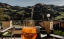 Ein Glas mit einem orangefarbenen Getränk steht auf einer Terrasse mit Blick auf grüne Berge. Die Atmosphäre ist entspannt und einladend.