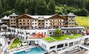 A luxurious hotel building surrounded by trees and green landscape. In the foreground, there is a pool area with loungers and a well-kept garden area.
