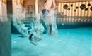 An indoor pool with two people jumping into the water. Splashes and waves occur as they dive into the cool water.