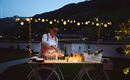 A woman is preparing drinks at a decorated cart at night. The surroundings are adorned with lights and offer a beautiful view of the landscape.