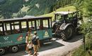 A family is standing by a path in front of a tractor and a cart in a picturesque mountainous landscape. In the background, green hills and a clear blue sky can be seen.