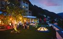 A beautiful garden with illuminated pavilions and an inviting atmosphere. In the background, mountains and a clear evening sky can be seen.