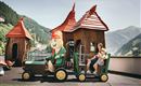 A playground with colorful playhouses and a small train attraction. A cheerful child is sitting next to a mascot and enjoying time outdoors.