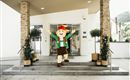 A cheerful mascot figure stands in front of a building and greets visitors. Surrounded by plants and an inviting atmosphere, it appears friendly and welcoming.