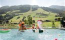 A family is having fun in the pool with an airy unicorn swing. In the background, green hills and traditional houses can be seen.
