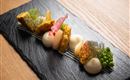 An artistically arranged dish on a black stone. It contains fried pieces, creamy side dishes, and fresh vegetables.