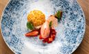 An artfully arranged dessert on a blue plate. It contains a yellow cream, strawberries, and a pink sorbet.
