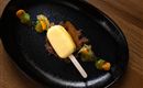 An artistically arranged dessert on a black plate. It consists of a creamy popsicle and colorful garnishes.