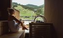 A woman is sitting relaxed in a sauna and enjoying the view of green hills. The atmosphere is calm and inviting.