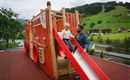 A child happily slides down a slide of a colorful play structure. In the background, green hills and a beautiful landscape can be seen.