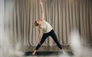 A woman is performing a yoga exercise in a bright room. The room has a neutral background with curtains.
