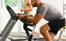 A man is training on a stationary bike while a woman jogs on a treadmill in the background. The room has large windows that let in natural light.