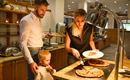 A family is enjoying a meal in a restaurant. The woman is serving pizza while the man and his son are waiting.