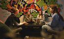 Four women are sitting around a table playing cards. In the background, there is a colorful wall covered in graffiti.