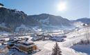 Eine schneebedeckte Berglandschaft mit einem kleinen Dorf und vielen Häusern. Die Sonne scheint über die Berge und die Skigebiete sind sichtbar.