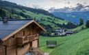 Ein schönes Holzhaus in einer grünen Hügellandschaft. Im Hintergrund sind Berge und kleine Dörfer zu sehen.
