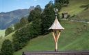 Ein traditioneller Dachreiter mit einer Wetterfahne, der auf einem modernen Gebäude steht. Im Hintergrund sind sanfte Hügel und Bäume vor einer Bergkulisse zu sehen.
