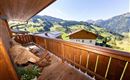 A beautiful balcony with a wooden railing and a view of green hills and mountains. Relaxed seating area decorated with a blanket and plants.
