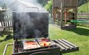 Ein Grill steht im Grünen und raucht, während Würstchen und Gemüse darauf garen. Im Hintergrund sieht man einen Spielplatz und eine malerische Landschaft.