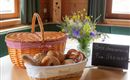 Ein Korb mit frischen Brötchen und Brezeln steht auf einem Tisch. Daneben steht ein Blumenstrauß und eine Tafel mit dem Hinweis "Brötchenservice Fam. Steiner".