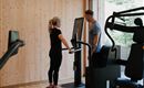 A woman stands at a fitness machine while a man gives her instructions. The room has wooden walls and large windows that let in a lot of light.