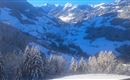 A winter mountain landscape with snow-covered trees and a clear, blue sky. In the background, majestic mountains and a snowy valley can be seen.