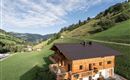 Ein Holzhaus umgeben von grünen Wiesen und sanften Hügeln. Der Himmel ist klar mit wenigen Wolken.