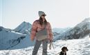 A woman stands in the snow with her dog. In the background, you can see snow-capped mountains and a clear sky.