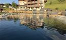 A beautiful hotel by the water, surrounded by mountains and green grass. The sky is blue and the atmosphere is calm.