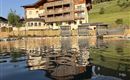 A beautiful hotel by the water with a wood-paneled facade. In the background, lush green hills and a blue sky can be seen.