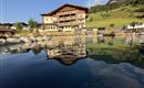 A beautiful hotel with a wooden facade is located by the water. The surrounding landscape is green and mountainous.