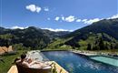 A relaxing panorama with a pool and a wooden terrace. In the background, green mountains and a clear blue sky can be seen.