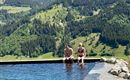 A floating infinity pool with a view of green mountains. Two people are sitting at the edge of the pool and enjoying the view.