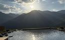A tranquil pool extending before majestic mountains, under a clear sky with radiant sun. The gentle waves of the water reflect the surrounding landscape.