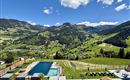 A beautiful landscape with green hills and mountains. In the foreground, there is a pool surrounded by loungers.