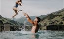 A father lifts his child into the air in the pool. In the background, green hills and a serene landscape can be seen.