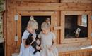 Two girls are standing in front of a rabbit hutch holding a black rabbit. They are wearing light dresses and smiling kindly.
