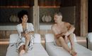 A couple sits relaxed on loungers in a stylish room. They smile at each other and enjoy their time together.