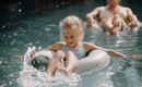 A smiling girl is sitting in an inflatable ring in the water. In the background, another person can be seen in the pool.