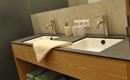 A modern bathroom with a wooden washbasin and two sinks. The walls are painted in neutral colors, and a towel is placed next to the sinks.