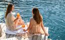 Zwei Frauen sitzen am Rand eines Pools und genießen Cocktails. Das Wasser glitzert und es ist eine entspannte Atmosphäre.