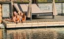 Zwei Frauen liegen entspannt auf einer Holzplattform am Wasser und genießen Cocktails. Im Hintergrund ist ein Schattenplatz mit Liegestühlen zu sehen.