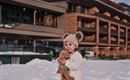 Ein kleines Kind steht im Schnee und hält ein Teddybär. Im Hintergrund ist ein modernes Gebäude zu sehen.