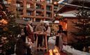 Eine Gruppe von Menschen steht um einen Tisch in einem winterlichen Außenbereich. Im Hintergrund sieht man ein Holzhaus und Weihnachtslichter, während ein Feuer knistert.