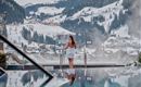 Eine Frau in einem Bademantel steht am Rand eines Pools und blickt auf schneebedeckte Berge. Der Pool spiegelt die Landschaft wider und schafft eine entspannende Atmosphäre.