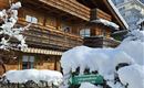 A cozy wooden house in the snow with a balcony and snow-covered trees. A sign points the way to a vacation apartment.