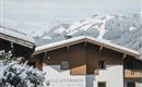 A cozy house with a wooden façade in a snowy mountain landscape. In the background, majestic mountains and a clear sky can be seen.