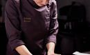 Eine Bäckerin formt mit ihren Händen eine Teigmasse in der Küche. Auf dem Tisch liegt ein Stück Biskuit und eine Schüssel mit frischen Erdbeeren.