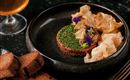 Ein appetitlicher Teller mit Tatar, garniert mit bunten Kräutern und essbaren Blüten. Dazu knusprige Chips und Scheiben von dunklem Brot.