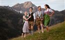 Eine Gruppe von vier Personen in traditioneller Tracht steht auf einer Wiese mit Bergen im Hintergrund. Sie lächeln und wirken fröhlich in der Natur.